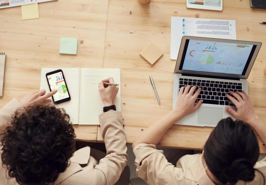 Two people at a wooden table, one writing in a notebook and one typing on a laptop with charts