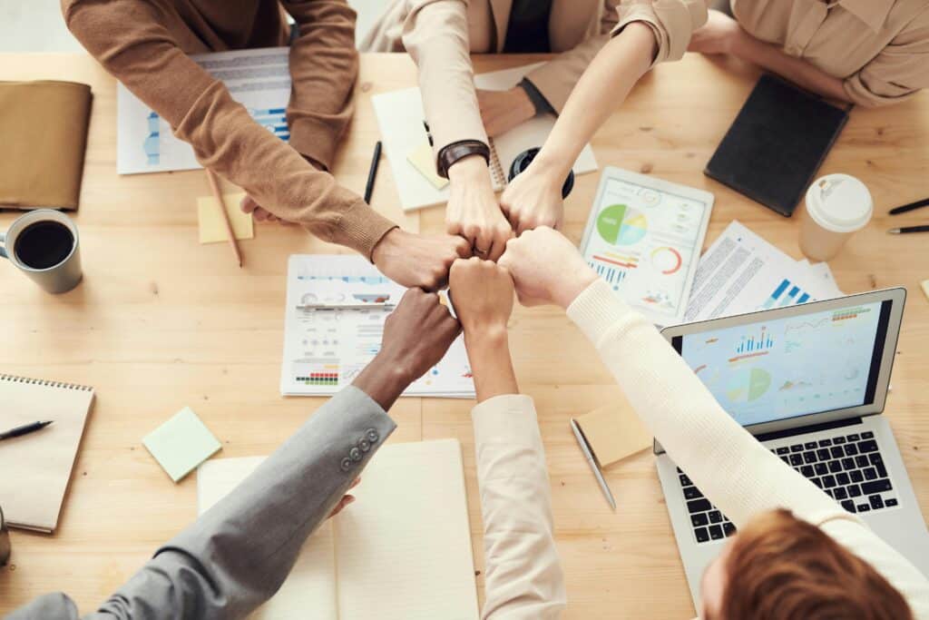 Hands joined in a fist bump above a table with charts, a laptop, notebooks and coffee