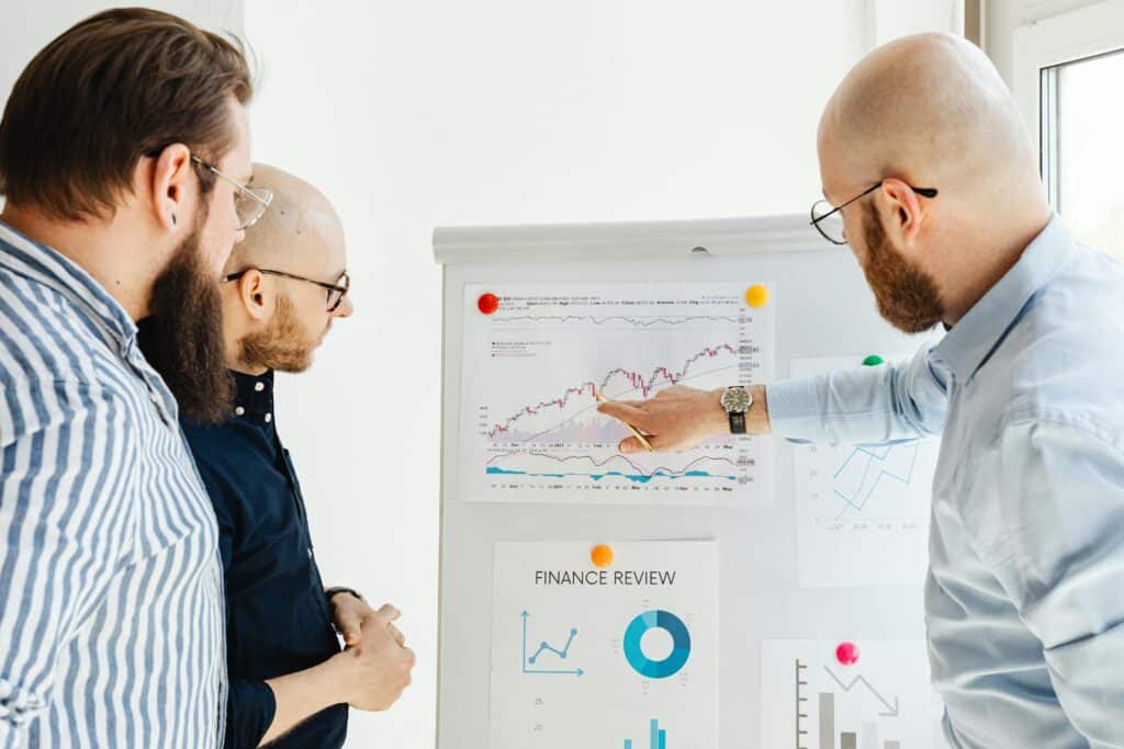 Three bearded men looking at a flipchart with line graphs, one of them pointing at it