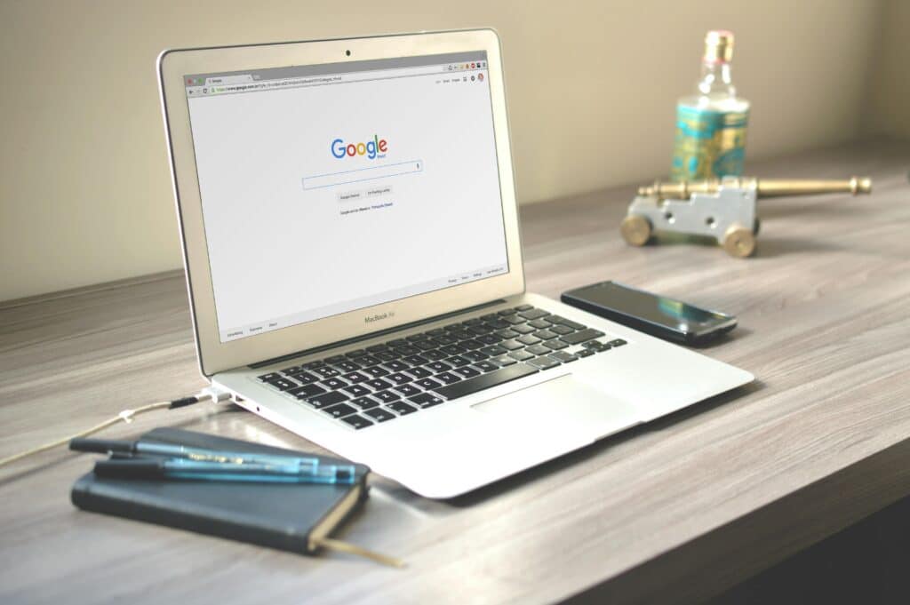 Laptop showing the Google search page on a wooden desk with a notebook, pens and a phone
