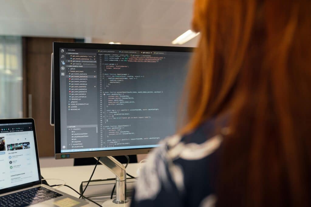 Rudowłosa osoba widziana od tyłu patrzy na monitor pełen kodu obok laptopa