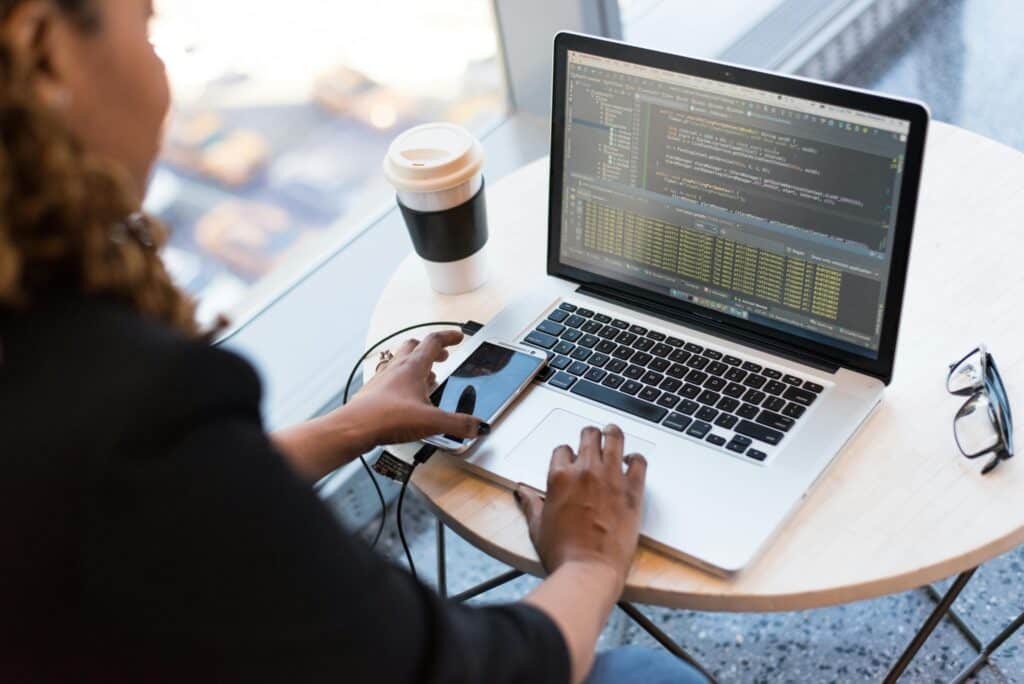 Woman typing on a laptop showing code, with a phone, takeaway coffee cup and glasses on a round table