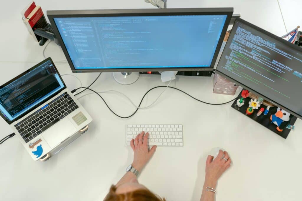 Desk seen from above with two monitors and a laptop showing code, hands on a keyboard and mouse
