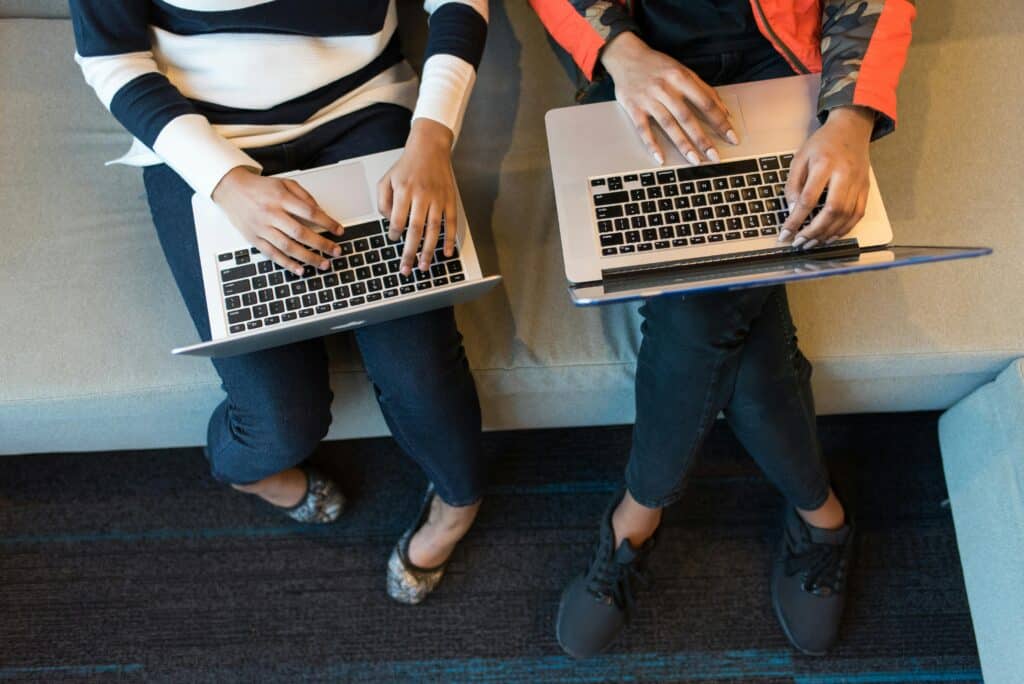 Two people sitting side by side on a sofa with laptops on their knees, seen from above